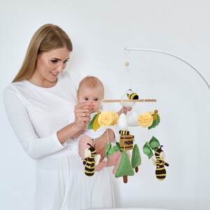 May include: A white crib with a white and yellow baby mobile hanging above it. The mobile features bees, flowers, and a honey pot. A woman is holding a baby in the crib.
