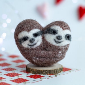 May include: Two brown and white felt sloths hugging in the shape of a heart. The sloths are sitting on a small wooden base.