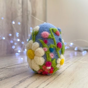 Könnte beinhalten: Ein blaues Filz-Osterei, verziert mit weißen, gelben, rosa und grünen Filzblumen. Das Ei liegt auf einer hellbraunen Holzoberfläche.