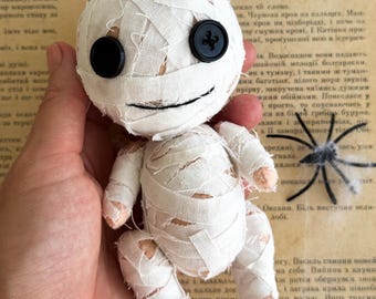 May include: A handmade mummy doll with a round head, wrapped in white fabric bandages. It has two large black button eyes and a stitched smile. The doll is held in a hand, with an open book in the background.