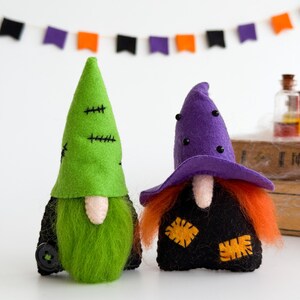 May include: Two Halloween gnome figures. One gnome has a green hat with black stitching and a green beard. The other gnome has a purple witch hat, orange hair, and a black outfit with yellow patches. A banner of orange, purple, and black flags hangs in the background.