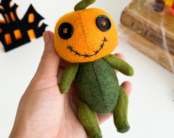 May include: A handmade Halloween pumpkin doll with an orange head, black button eyes, and a stitched smile. The body is green, and the doll is held in a hand. A black haunted house decoration is in the background.