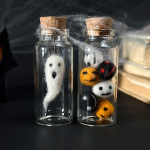 以下が含まれることがあります： コルク栓付きの小さなガラス瓶が2つ。1つには白いフェルトの幽霊、もう1つにはフェルトのかぼちゃ、幽霊、黒いボールが入っています。ハロウィーンをテーマにした装飾が、クモの巣のある暗い表面に展示されています。