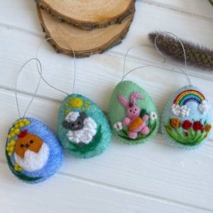 Puede incluir: Cuatro adornos de huevos de Pascua de fieltro con diferentes diseños. Un huevo es azul con un pollito amarillo dentro, otro es verde con una oveja blanca, otro es verde con un conejito rosa y el último es azul con un arcoíris y flores.