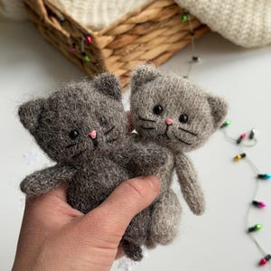 K&ouml;nnte beinhalten: Zwei handgefertigte graue Katzenpl&uuml;schtiere mit rosa Nasen und schwarzen Schnurrhaaren werden in einer Hand gehalten. Die Katzen haben kleine Ohren und bestehen aus einem flauschigen Material. Der Hintergrund umfasst einen Weidenkorb und Lichterketten.