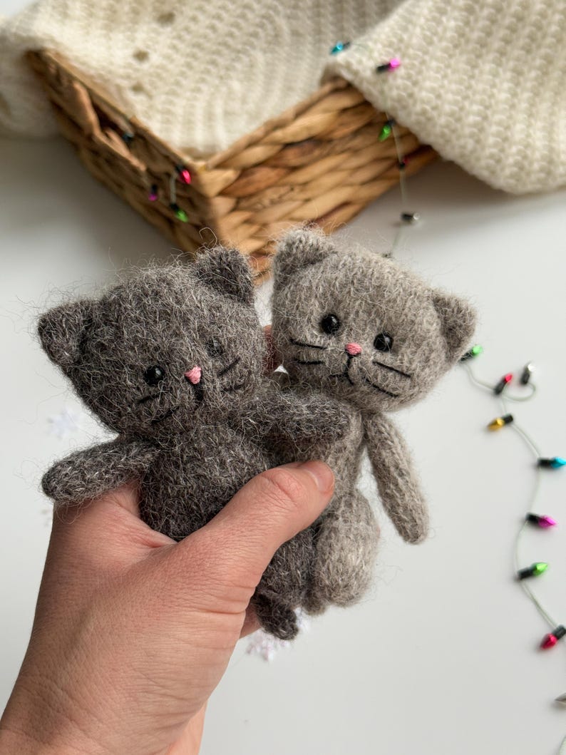 K&ouml;nnte beinhalten: Zwei handgefertigte graue Katzenpl&uuml;schtiere mit rosa Nasen und schwarzen Schnurrhaaren werden in einer Hand gehalten. Die Katzen haben kleine Ohren und bestehen aus einem flauschigen Material. Der Hintergrund umfasst einen Weidenkorb und Lichterketten.