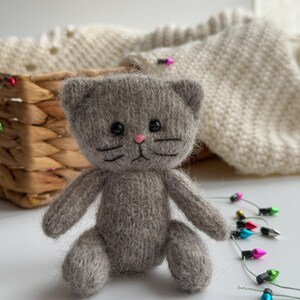 K&ouml;nnte beinhalten: Eine handgefertigte, graue, gestrickte Katzendoll mit schwarzen, gestickten Schnurrhaaren und Augen sowie einer rosa Nase. Die Katze sitzt mit ausgestreckten Armen und Beinen. Ein Weidenkorb und ein cremefarbener Strickpullover befinden sich im Hintergrund. Bunte Weihnachtslichter sind im Vordergrund.