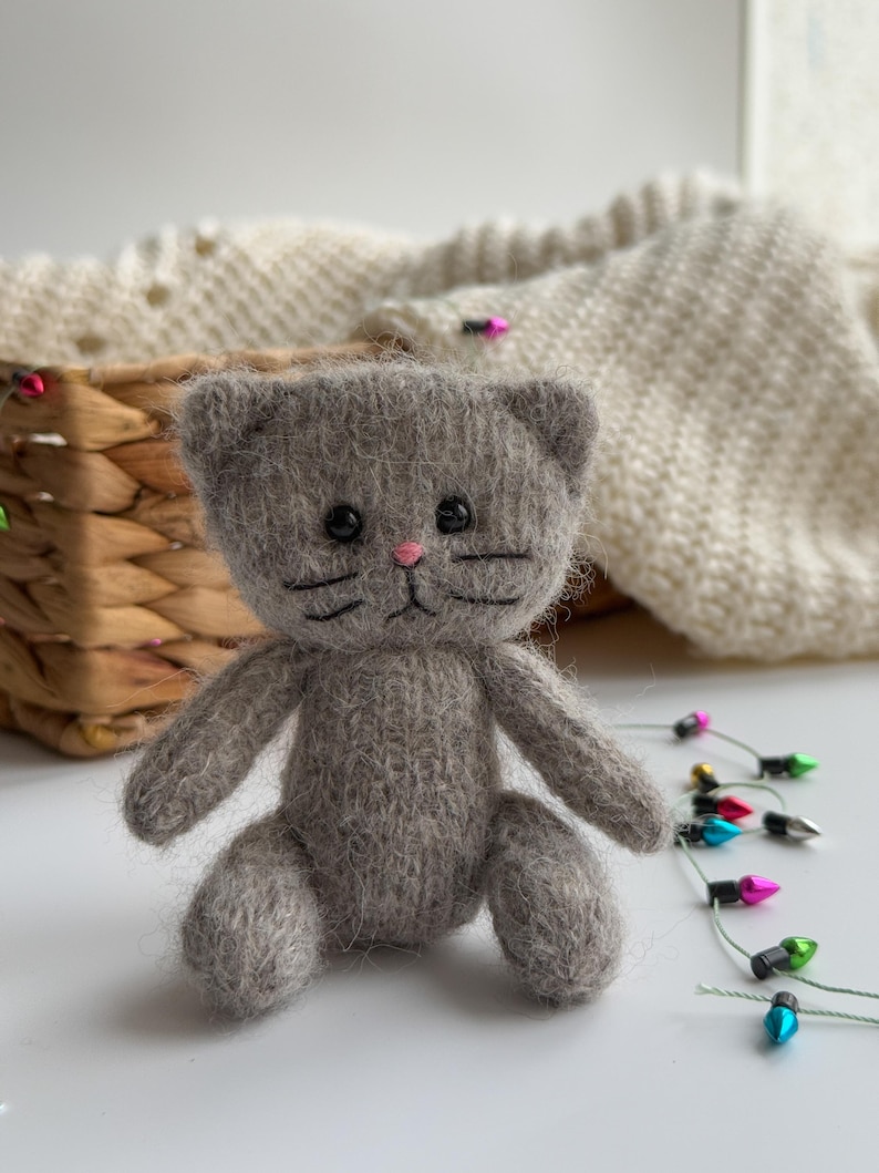 K&ouml;nnte beinhalten: Eine handgefertigte, graue, gestrickte Katzendoll mit schwarzen, gestickten Schnurrhaaren und Augen sowie einer rosa Nase. Die Katze sitzt mit ausgestreckten Armen und Beinen. Ein Weidenkorb und ein cremefarbener Strickpullover befinden sich im Hintergrund. Bunte Weihnachtslichter sind im Vordergrund.
