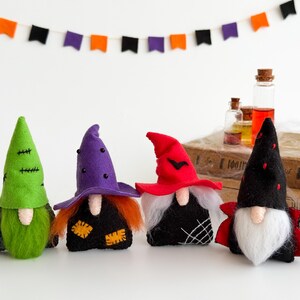 May include: Four Halloween gnome figurines with pointed hats in green, purple, and red, and black outfits. Each gnome has a white beard and a unique design. A banner of orange, purple, and black flags hangs above them.