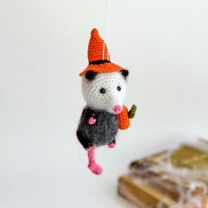 May include: A crocheted opossum wearing a witch hat and holding a small orange pumpkin. The opossum is hanging from a branch and has pink feet.
