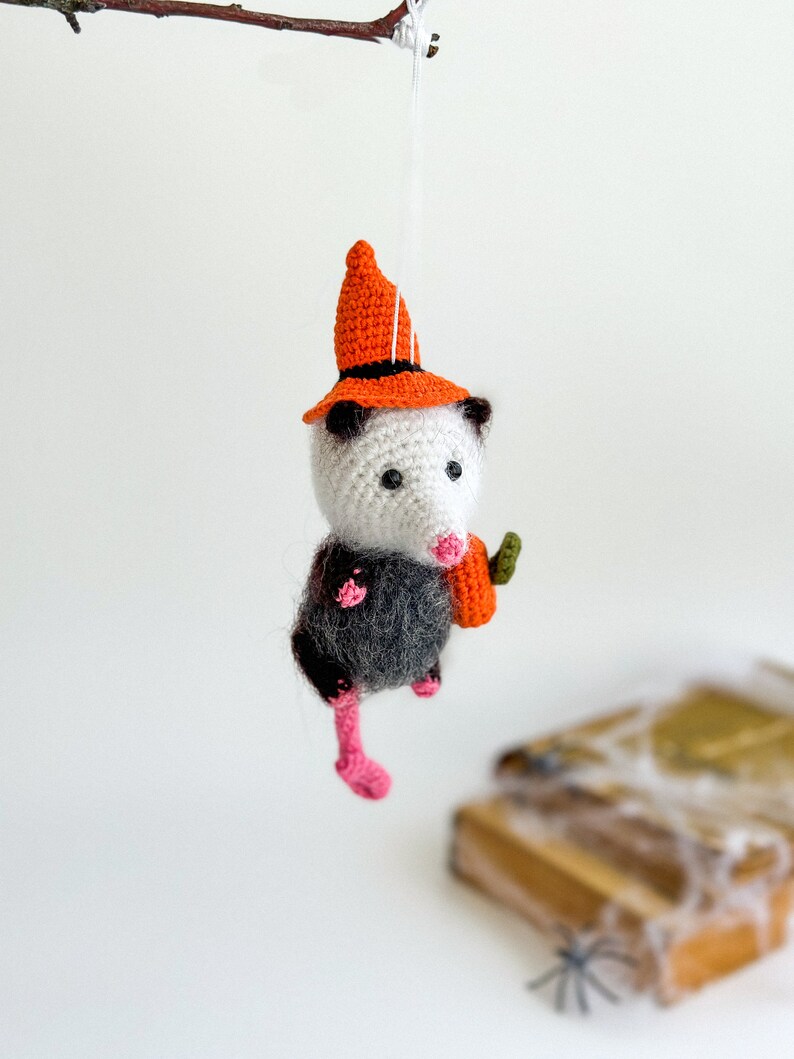 May include: A crocheted opossum wearing a witch hat and holding a small orange pumpkin. The opossum is hanging from a branch and has pink feet.