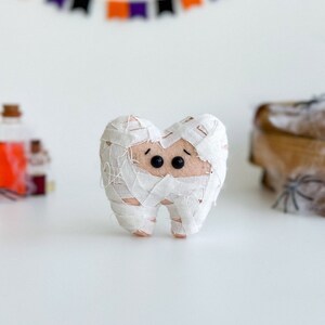 May include: A felt tooth wrapped in white bandages, resembling a mummy, with black button eyes. A Halloween-themed decoration. A banner with orange, purple, and black flags is in the background.