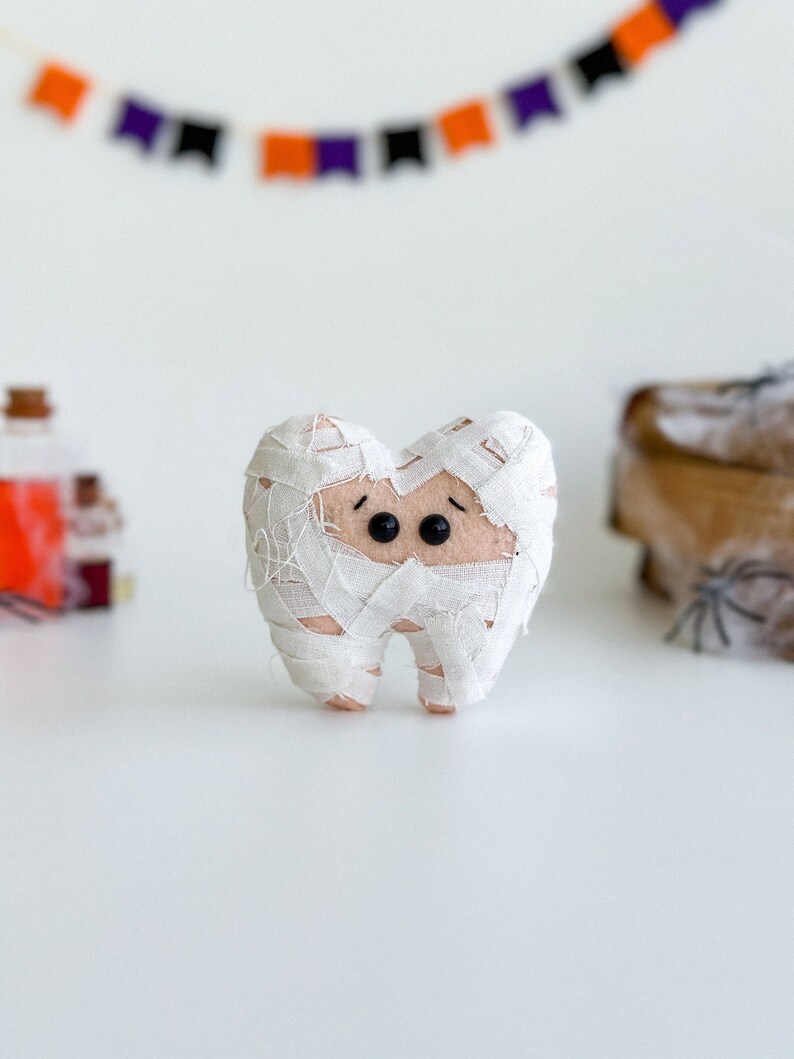 May include: A felt tooth wrapped in white bandages, resembling a mummy, with black button eyes. A Halloween-themed decoration. A banner with orange, purple, and black flags is in the background.