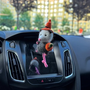 May include: A crocheted opossum wearing a witch hat and holding a small orange pumpkin. The opossum is hanging from the rearview mirror of a car.