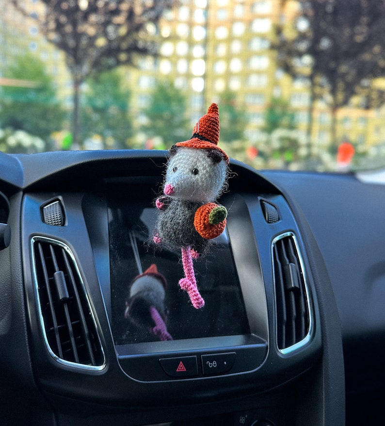May include: A crocheted opossum wearing a witch hat and holding a small orange pumpkin. The opossum is hanging from the rearview mirror of a car.