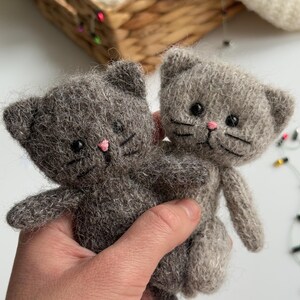 K&ouml;nnte beinhalten: Zwei graue, gestrickte Katzen-Stofftiere mit rosa Nasen und schwarzen Augen. Die Katzen haben kleine Ohren und Schnurrhaare. Die Katzen werden in der Hand einer Person gehalten.