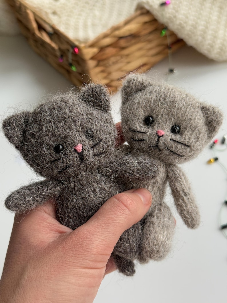 K&ouml;nnte beinhalten: Zwei graue, gestrickte Katzen-Stofftiere mit rosa Nasen und schwarzen Augen. Die Katzen haben kleine Ohren und Schnurrhaare. Die Katzen werden in der Hand einer Person gehalten.
