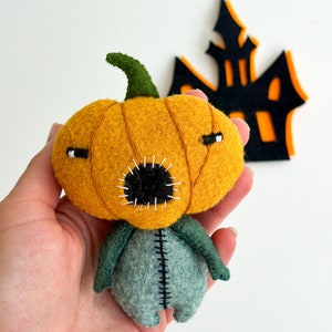 May include: A felt pumpkin character with a stitched mouth and eyes. The pumpkin is orange and has a green stem. The character has a green and grey body with stitched seams.