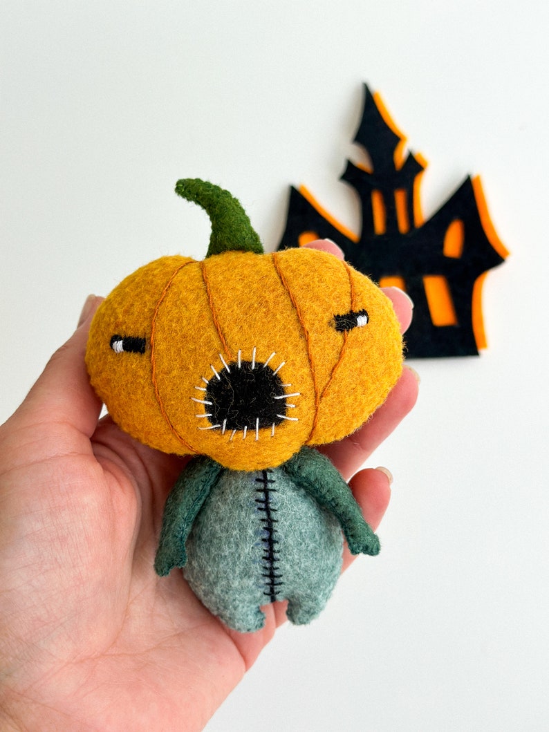 May include: A felt pumpkin character with a stitched mouth and eyes. The pumpkin is orange and has a green stem. The character has a green and grey body with stitched seams.