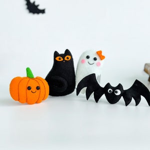 May include: A set of four felt Halloween decorations: a black cat, a white ghost with a pink bow, a black bat, and an orange pumpkin with a smiling face.