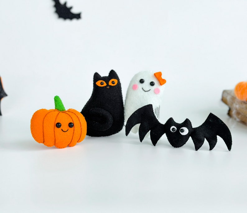 May include: A set of four felt Halloween decorations: a black cat, a white ghost with a pink bow, a black bat, and an orange pumpkin with a smiling face.