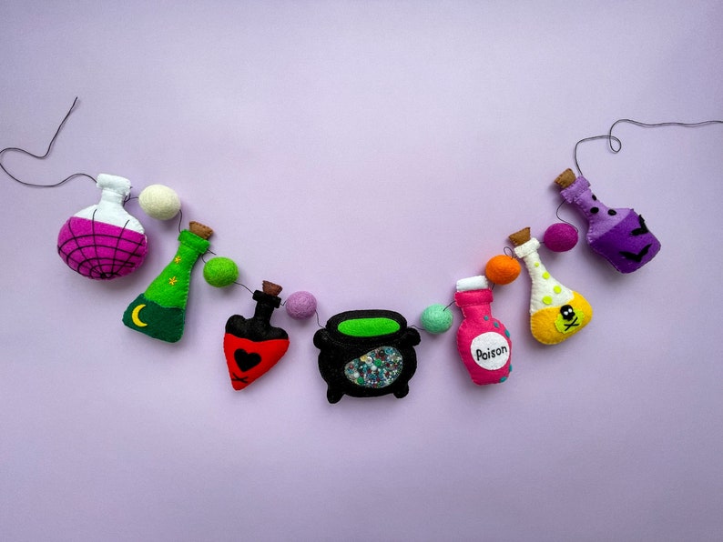 K&ouml;nnte beinhalten: Eine Halloween-Girlande mit Filz-Tr&auml;nkflaschen, einem Kessel und einer herzf&ouml;rmigen Flasche. Die Girlande ist mit wei&szlig;en Pompons und schwarzem Garn aufgef&auml;delt. Die Flaschen sind mit Sternen, einem Halbmond, einem Totenkopf und gekreuzten Knochen sowie dem Wort "Gift" verziert.