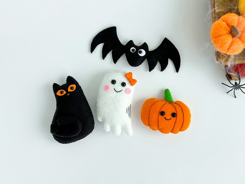 May include: A set of four felt Halloween decorations: a black cat, a white ghost, an orange pumpkin, and a black bat.