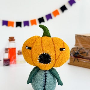 May include: A felt pumpkin character with a stitched-on mouth and eyes. The pumpkin is orange and the body is green. The character is standing on a white surface.