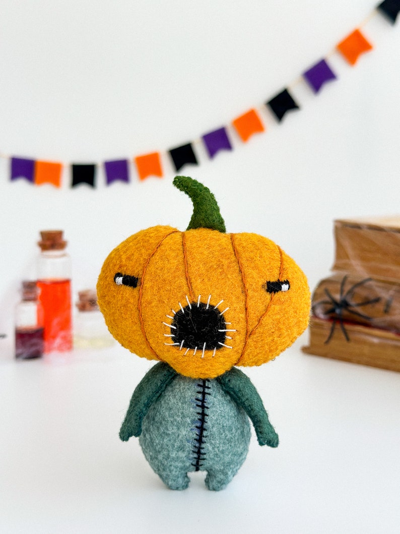 May include: A felt pumpkin character with a stitched-on mouth and eyes. The pumpkin is orange and the body is green. The character is standing on a white surface.