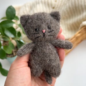 Könnte beinhalten: Ein handgefertigtes graues Katzenplüschtier mit rosa Nase und schwarzen Schnurrhaaren. Das kleine Stofftier hat ein schlichtes Design mit kleinen Ohren und ausgestreckten Armen. Das Plüschtier wird in der Hand einer Person gehalten.