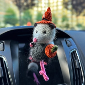 May include: A crocheted opossum wearing a witch hat and holding a small orange pumpkin. The opossum is a car charm with pink legs and a white face.
