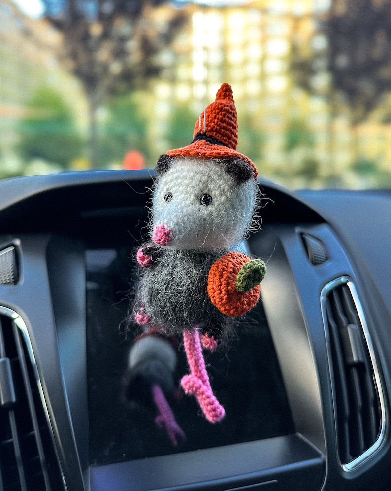 May include: A crocheted opossum wearing a witch hat and holding a small orange pumpkin. The opossum is a car charm with pink legs and a white face.