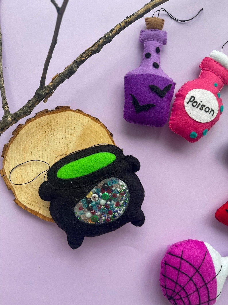Puede incluir: Un conjunto de adornos de Halloween de fieltro, que incluye un caldero lleno de cuentas de colores, una botella de poci&oacute;n morada con murci&eacute;lagos, una botella de poci&oacute;n rosa con la palabra "Poison" y un adorno de telara&ntilde;a rosa y negra.
