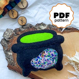 May include: A black felt cauldron with a green felt top and a colorful beaded design. The cauldron is sitting on a piece of wood with a "PDF pattern" label.