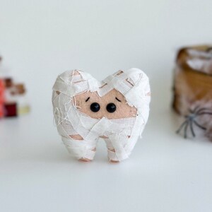 May include: A plush tooth-shaped toy wrapped in white fabric bandages, resembling a mummy. The toy has black button eyes and a light pink body. The background includes Halloween-themed decorations.