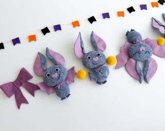 Könnte beinhalten: Eine Sammlung handgefertigter Halloween-Dekorationen aus Filz. Das Bild zeigt mehrere graue Fledermaus-Ornamente mit rosa Flügeln und herzförmigen Nasen, zusammen mit einer lila Schleife und einem Banner aus orangefarbenen, lila und schwarzen Flaggen. Gelbe Pompons sind ebenfalls enthalten.