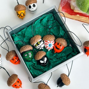 K&ouml;nnte beinhalten: Eine Sammlung von Halloween-Ackerln, die mit verschiedenen gruseligen Gesichtern bemalt sind, darunter ein Sch&auml;del, ein Geist, ein Zombie und ein K&uuml;rbis. Die Eicheln sind mit schwarzem Faden aufgereiht und in einer silbernen Schachtel mit gr&uuml;nem Seidenpapier ausgelegt.