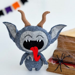 May include: A gray felt monster with horns, a wide open mouth, and a red tongue sticking out. The monster is standing on a white surface in front of a stack of books with a black spider web.