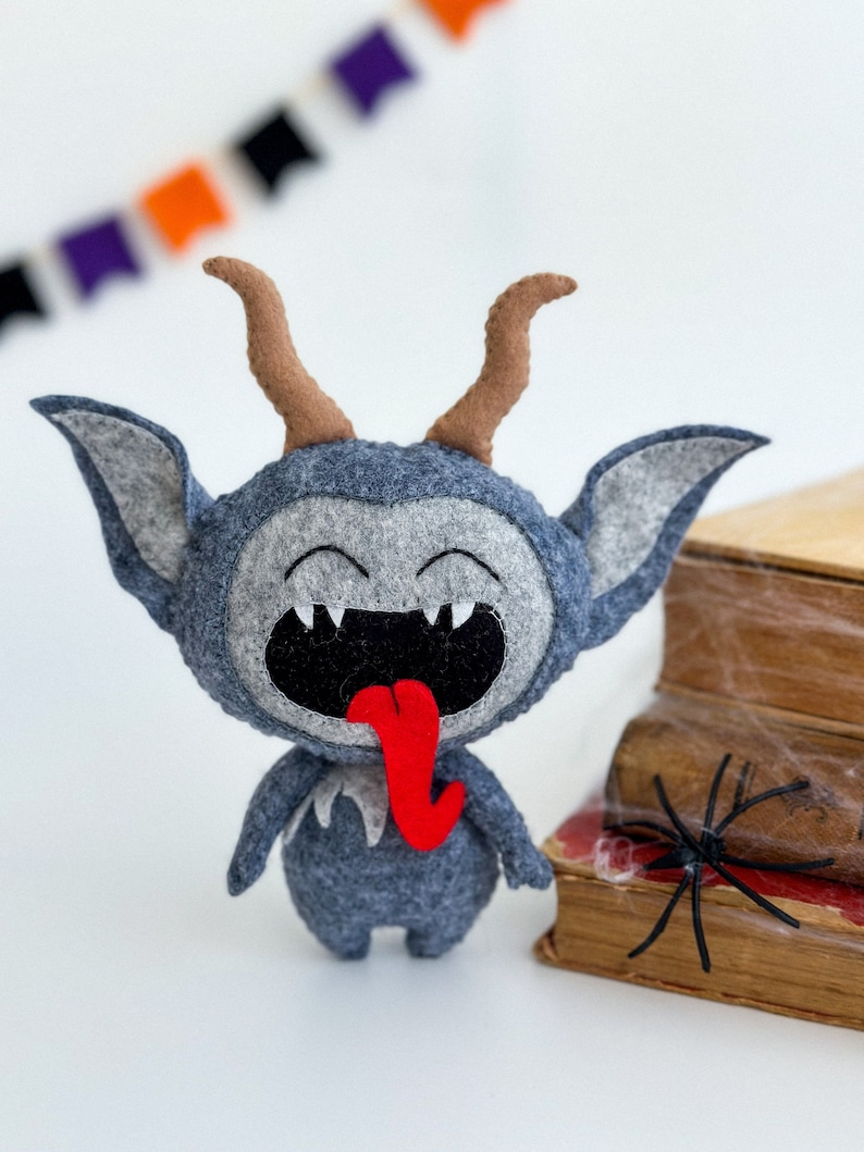 May include: A gray felt monster with horns, a wide open mouth, and a red tongue sticking out. The monster is standing on a white surface in front of a stack of books with a black spider web.