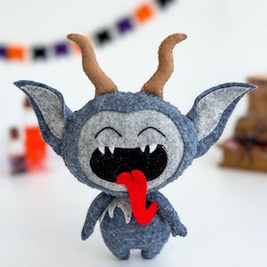 May include: A gray felt plush toy with brown horns and a red tongue sticking out. The toy has a wide, toothy grin and is a fun and whimsical Halloween decoration.