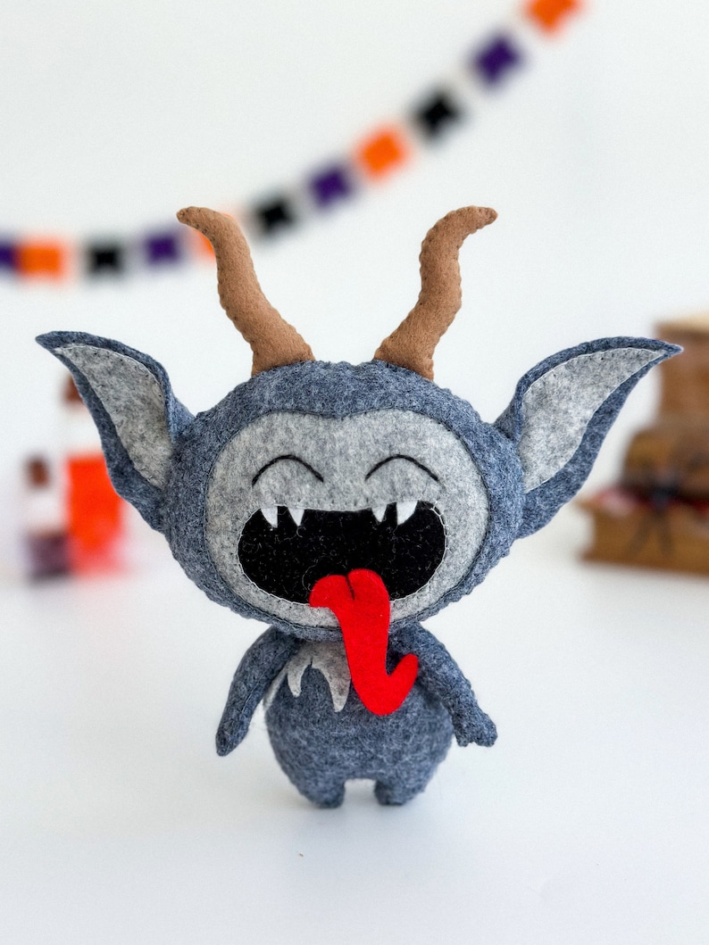 May include: A gray felt plush toy with brown horns and a red tongue sticking out. The toy has a wide, toothy grin and is a fun and whimsical Halloween decoration.