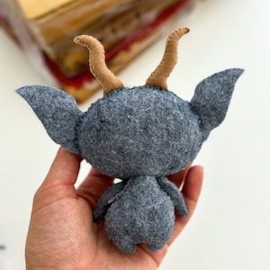 May include: A gray felt toy with brown felt horns. The toy is shaped like a small creature with a round head and a long, thin body. The toy is being held in a person's hand.
