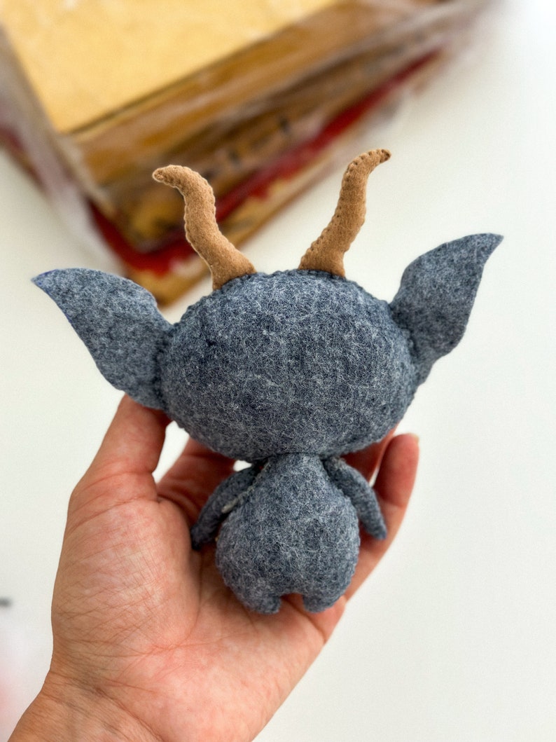 May include: A gray felt toy with brown felt horns. The toy is shaped like a small creature with a round head and a long, thin body. The toy is being held in a person's hand.