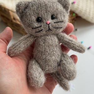 K&ouml;nnte beinhalten: Eine handgefertigte, graue, gestrickte Katzenpuppe mit schwarzen Augen, Schnurrhaaren und einer rosa Nase. Das kleine Pl&uuml;schtier hat ein detailliertes Design, einschlie&szlig;lich kleiner Ohren und eines Schwanzes. Die Katze wird in einer Hand gehalten, um ihre Gr&ouml;&szlig;e und Textur zu zeigen.