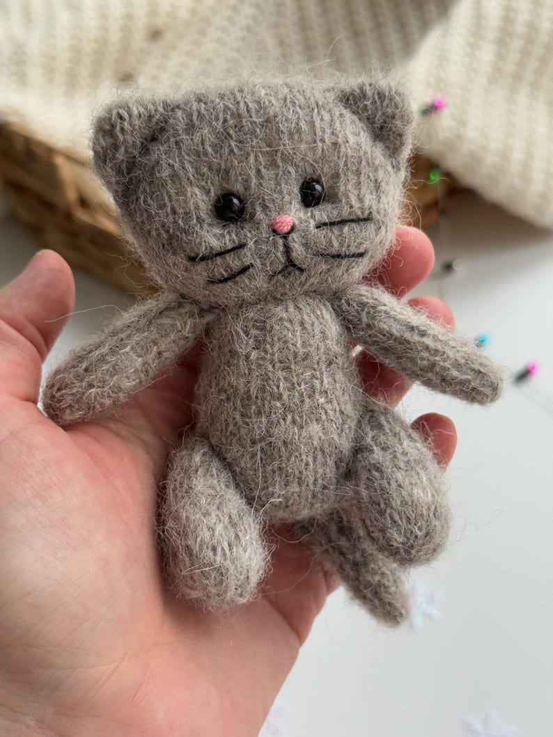 K&ouml;nnte beinhalten: Eine handgefertigte, graue, gestrickte Katzenpuppe mit schwarzen Augen, Schnurrhaaren und einer rosa Nase. Das kleine Pl&uuml;schtier hat ein detailliertes Design, einschlie&szlig;lich kleiner Ohren und eines Schwanzes. Die Katze wird in einer Hand gehalten, um ihre Gr&ouml;&szlig;e und Textur zu zeigen.