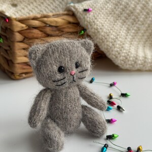K&ouml;nnte beinhalten: Eine kleine, graue, gestrickte Katzendoll mit schwarzen Augen, Schnurrhaaren und einer rosa Nase. Die Katze sitzt auf einer wei&szlig;en Oberfl&auml;che, mit einem geflochtenen Korb und einem cremefarbenen Strickartikel im Hintergrund. Eine Lichterkette mit bunten Weihnachtslichtern ist um die Katze drapiert.