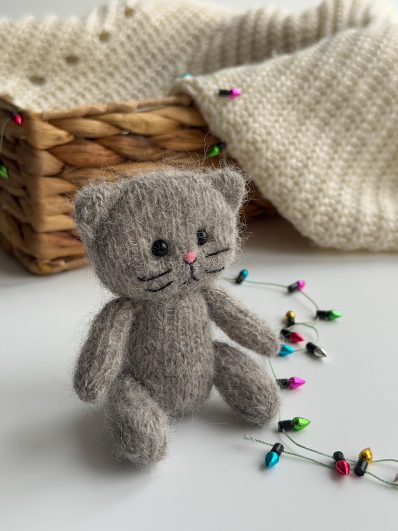 K&ouml;nnte beinhalten: Eine kleine, graue, gestrickte Katzendoll mit schwarzen Augen, Schnurrhaaren und einer rosa Nase. Die Katze sitzt auf einer wei&szlig;en Oberfl&auml;che, mit einem geflochtenen Korb und einem cremefarbenen Strickartikel im Hintergrund. Eine Lichterkette mit bunten Weihnachtslichtern ist um die Katze drapiert.