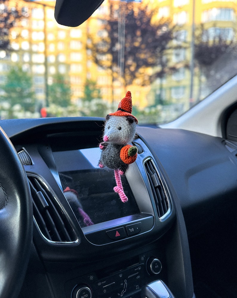 May include: A crocheted opossum wearing a witch hat and holding a small pumpkin. The opossum is hanging from the rearview mirror of a car.