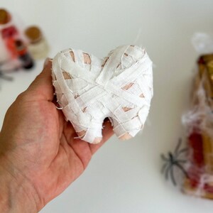 May include: A plush tooth wrapped in white bandages, resembling a mummy. The tooth is held in a hand against a white background, with blurred Halloween decorations in the background. A unique Halloween decoration.