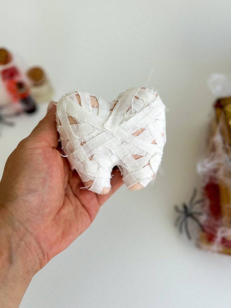 May include: A plush tooth wrapped in white bandages, resembling a mummy. The tooth is held in a hand against a white background, with blurred Halloween decorations in the background. A unique Halloween decoration.